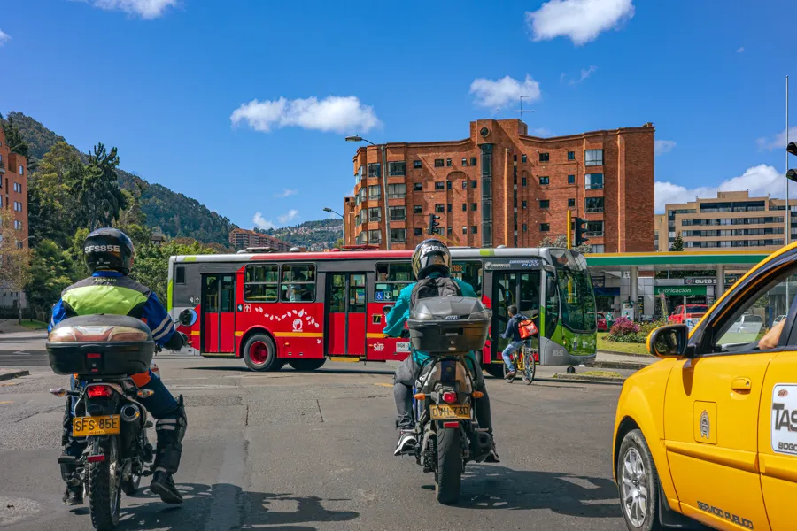 Un bus cruzando una intersección en Bogotá mientras dos motociclistas y un taxi amarillo esperan en la línea