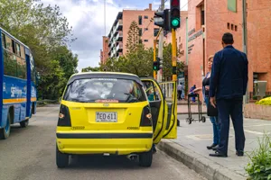 Un taxi amarillo detenido junto al andén con las puertas abiertas, justo debajo de un semáforo en verde en Bogotá, con un peatón esperando al lado