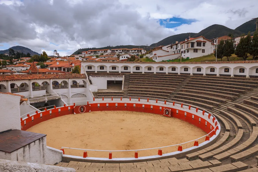 Guatavita's small bullring with a red barrier, tiered stone seating, and terracotta-roofed houses on the hillside behind