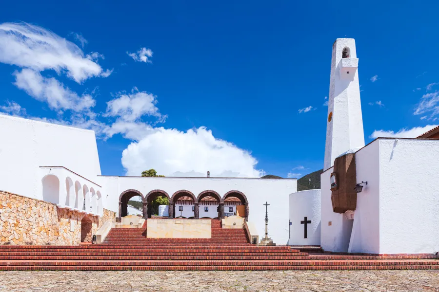 A whitewashed square in Guatavita with a tall bell tower, an arcaded gallery, and wide brick steps