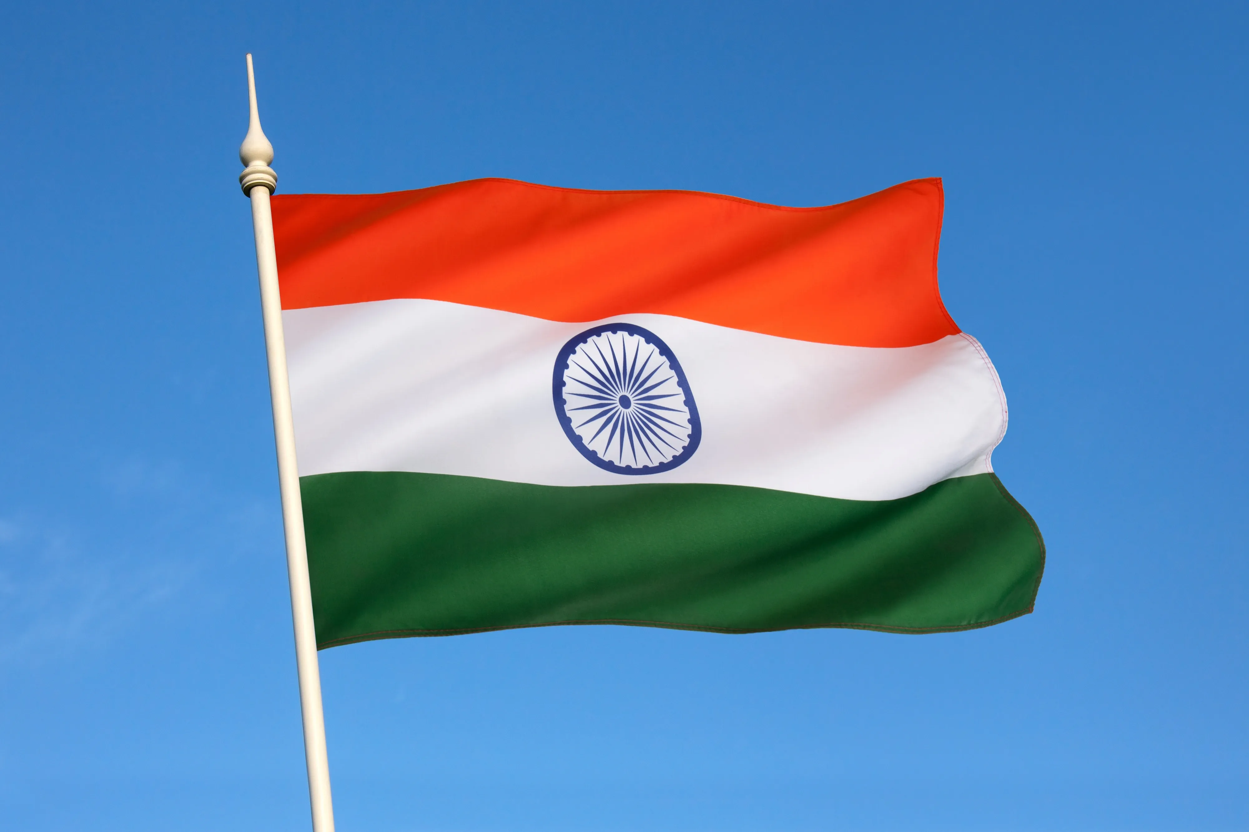 The Indian national flag, saffron, white, and green with a navy wheel at its centre, flying against a clear blue sky