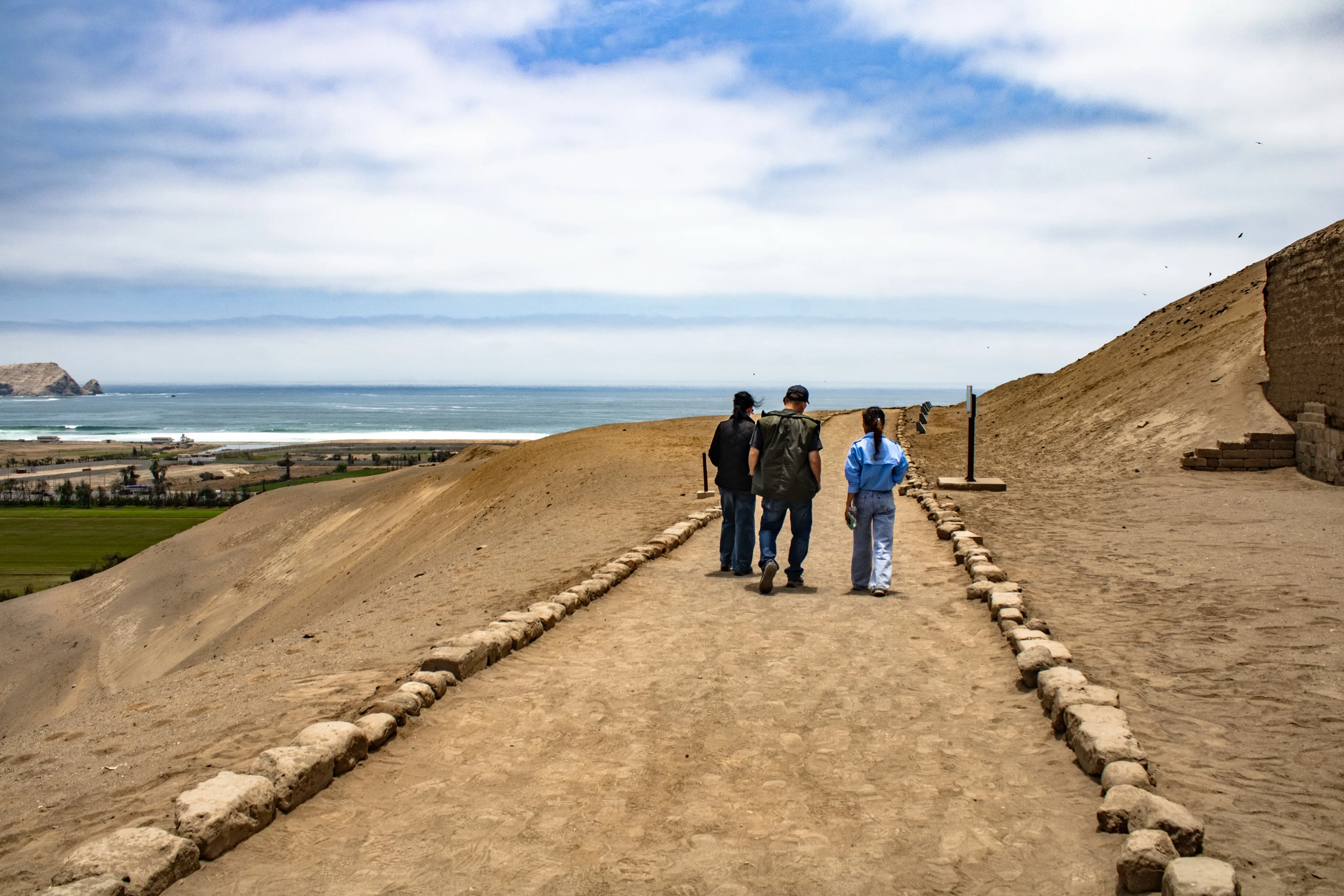 Visitantes caminando por un sendero en Pachacámac con el océano Pacífico a lo lejos