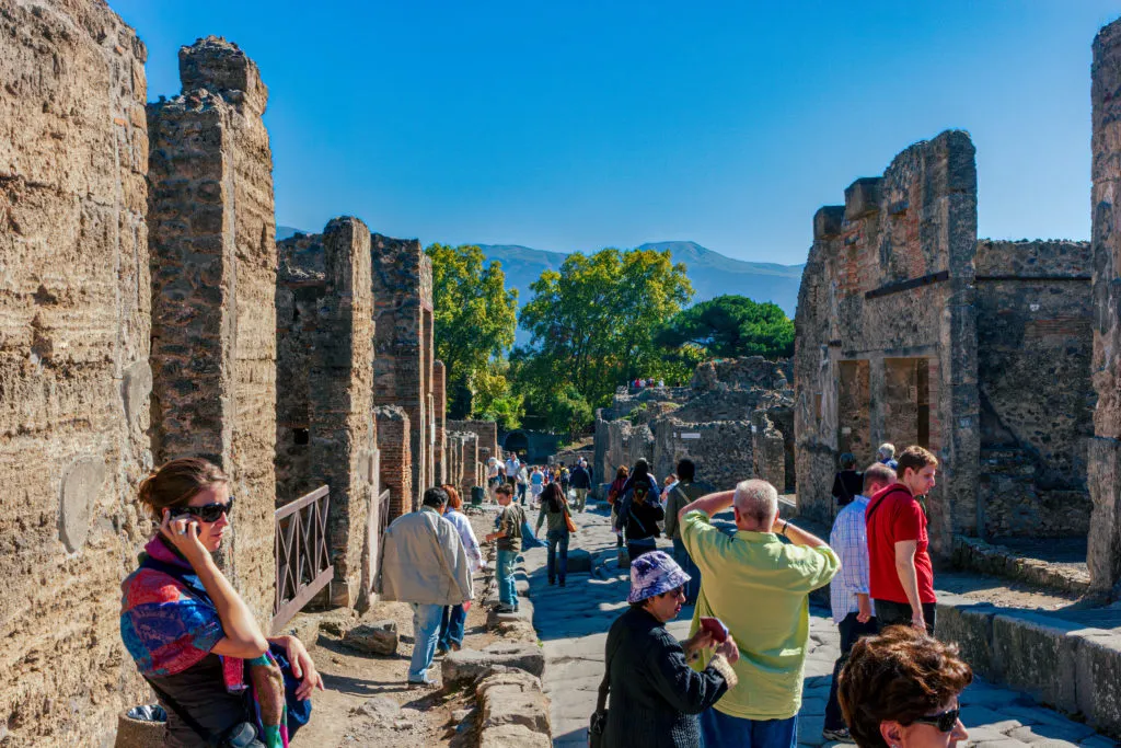 Visitors walking a paved street between the roofless stone walls of Pompeii, with the hills beyond