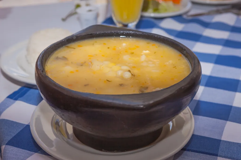 A clay bowl of ajiaco soup with corn and potato, served with rice and a glass of juice on a checked tablecloth